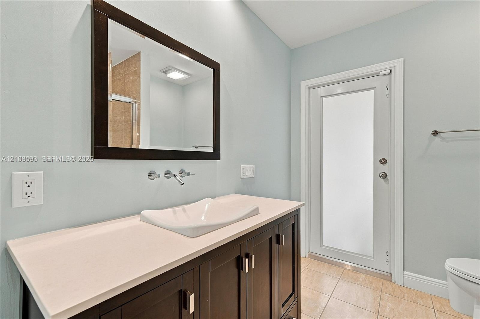 Vanity featuring a stone-surface countertop, rectangular vessel sink, wall-mounted faucet, framed wall mirror, and dark-finish cabinetry with metallic