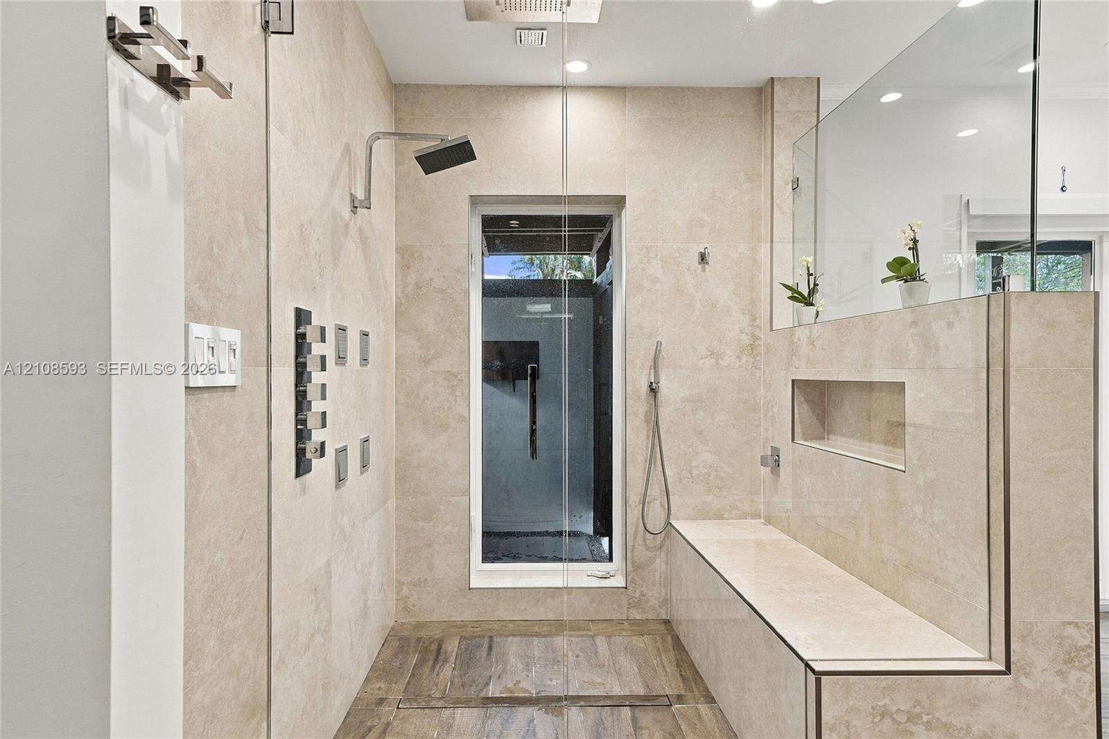 Walk-in shower featuring large-format neutral stone wall tile, multi-head shower system with a rain shower head, built-in stone bench with integrated 