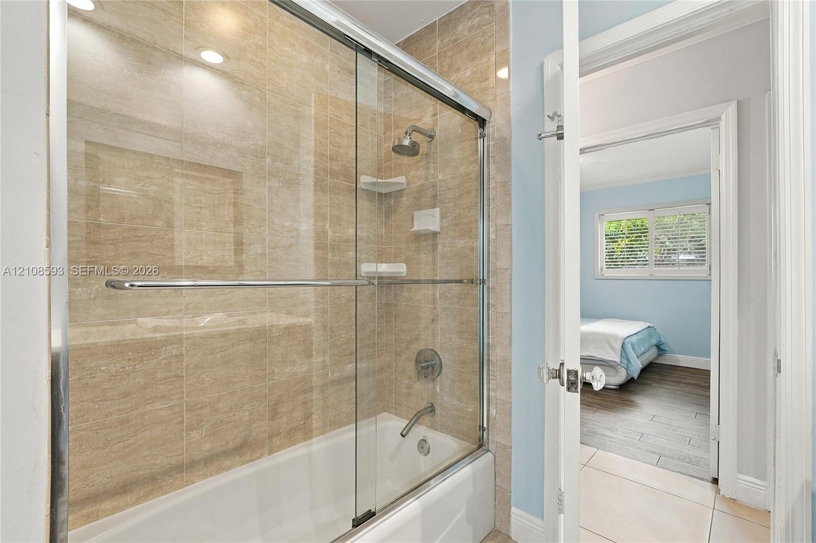 Bathtub and shower combination with floor-to-ceiling neutral tone tile surround, sliding glass enclosure, integrated corner shelving, and chrome fauce
