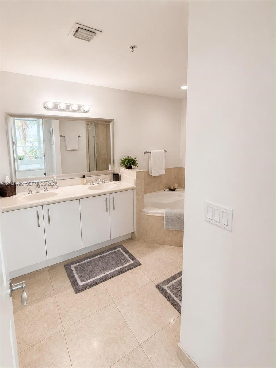 Double vanity with integrated sinks, neutral solid-surface countertop, full-width mirror, and tile flooring with a separate soaking tub featuring ston
