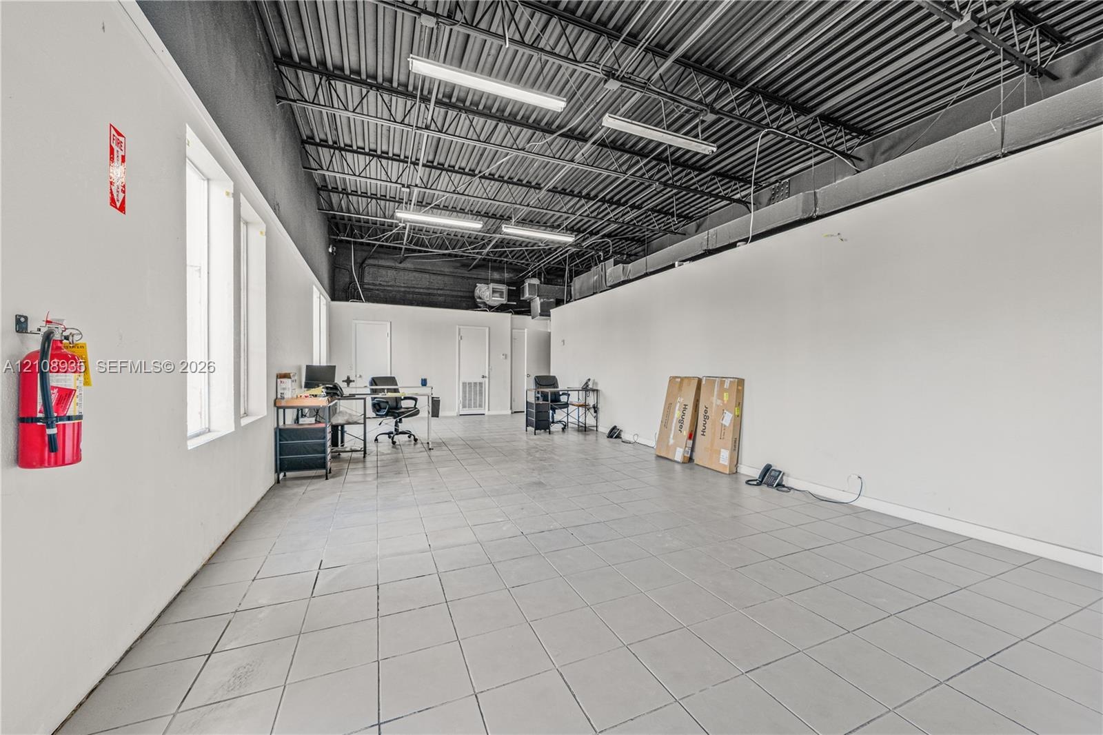 Expansive interior featuring high, exposed-joist ceilings, industrial-style black painted rafters, large rectangular ceramic tile flooring, and linear