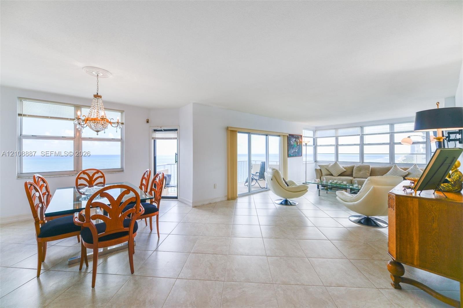 Open-concept main living area featuring light-toned tile flooring, oversized windows, multi-pane sliding glass doors, and a decorative crystal chandel