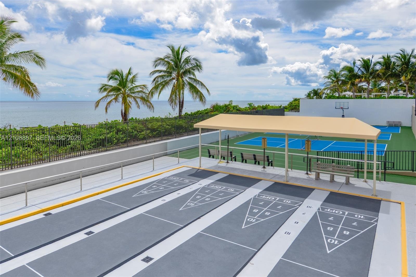 Shuffleboard courts with a shaded pavilion and a hard-surface sports court featuring a basketball hoop, set against an oceanfront backdrop with palm t