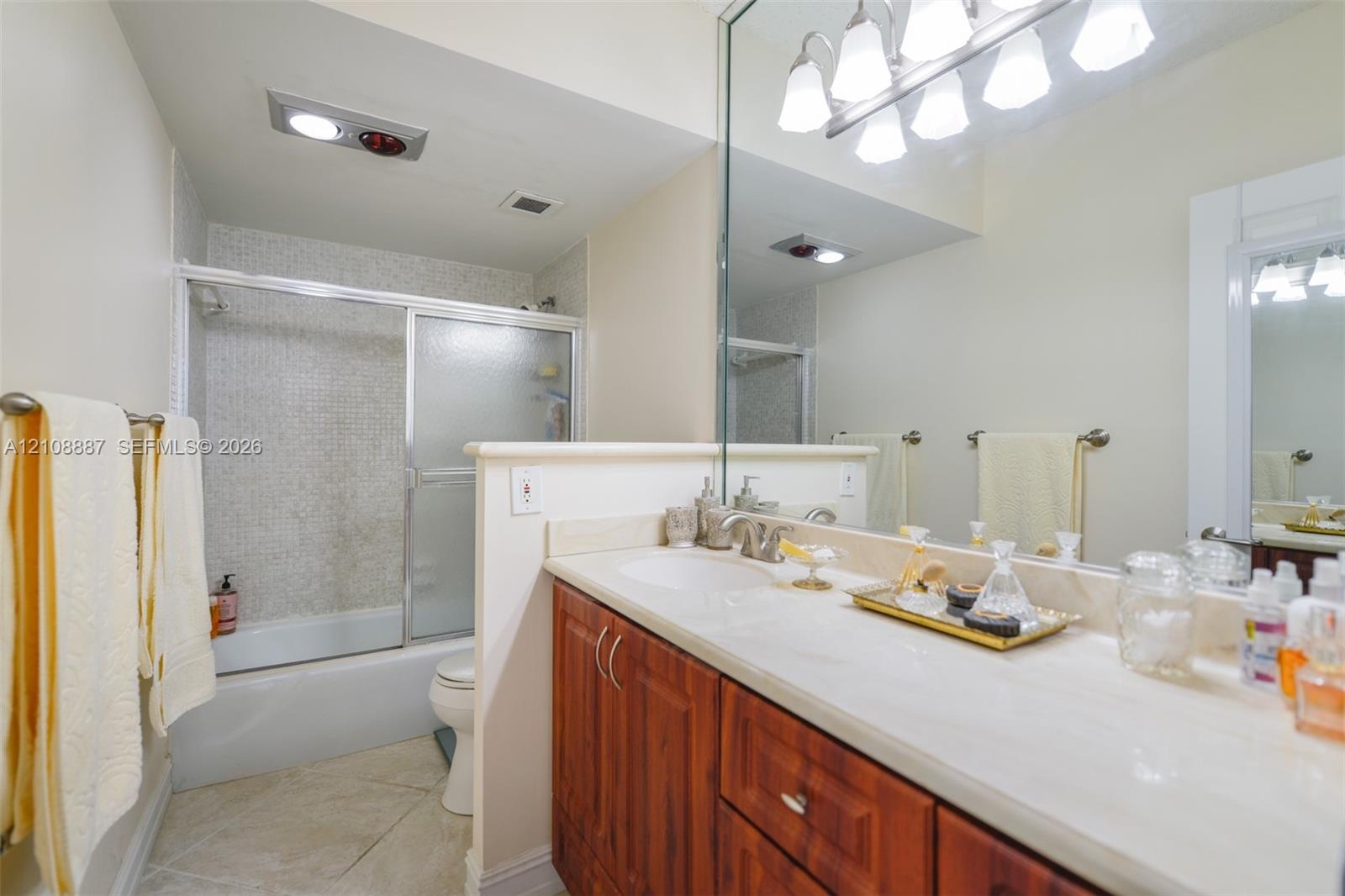Bathroom featuring a solid-surface countertop, wood-finish cabinetry, a full-width vanity mirror, a combination tub and shower with a sliding glass en