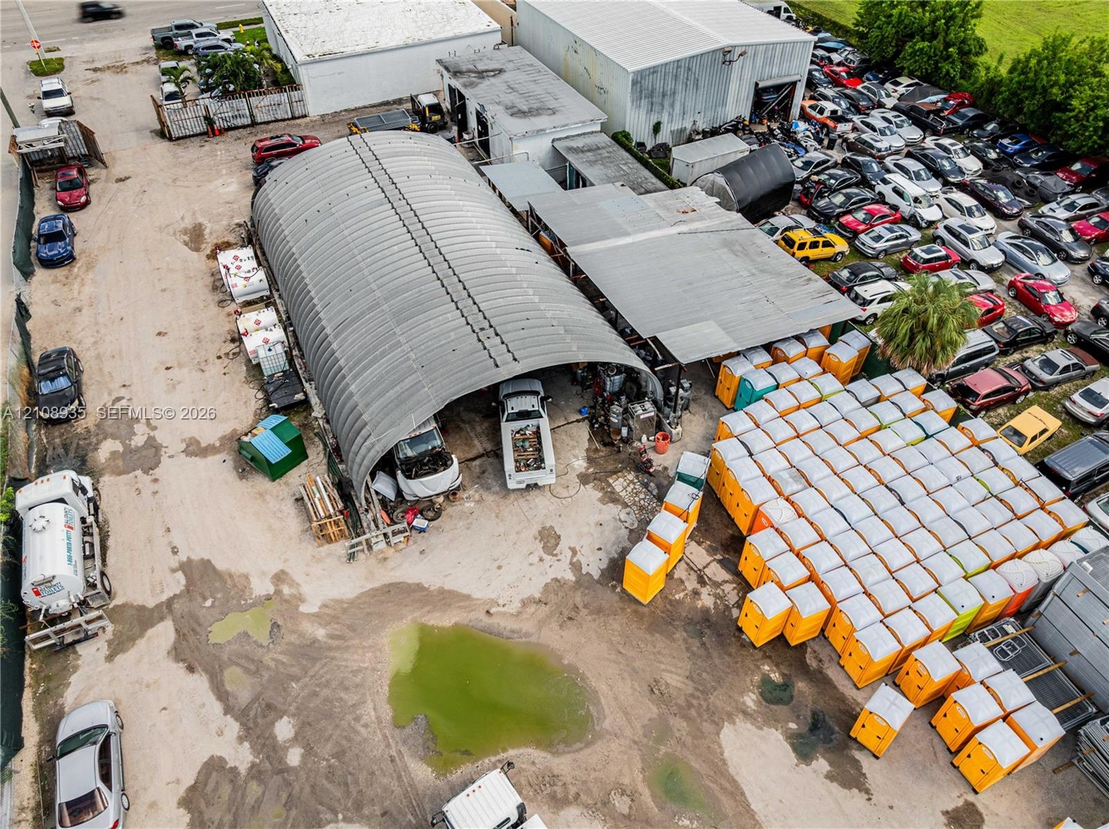 Industrial property featuring a metal arched structure and an expansive open-air parking lot with direct access to a paved roadway