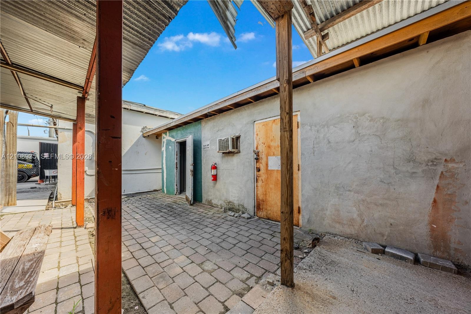 Covered outdoor area featuring paver flooring, exposed wooden support beams, and corrugated metal roofing