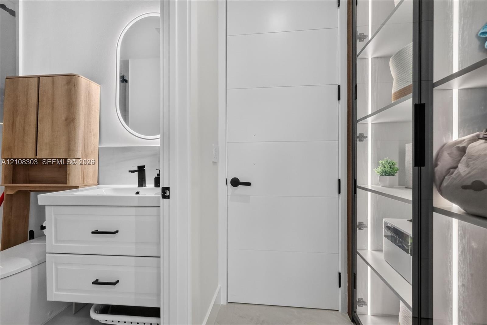 Bathroom featuring a white vanity with matte black hardware, integrated backlit oval mirror, wood-tone wall-mounted cabinet, and built-in shelving wit