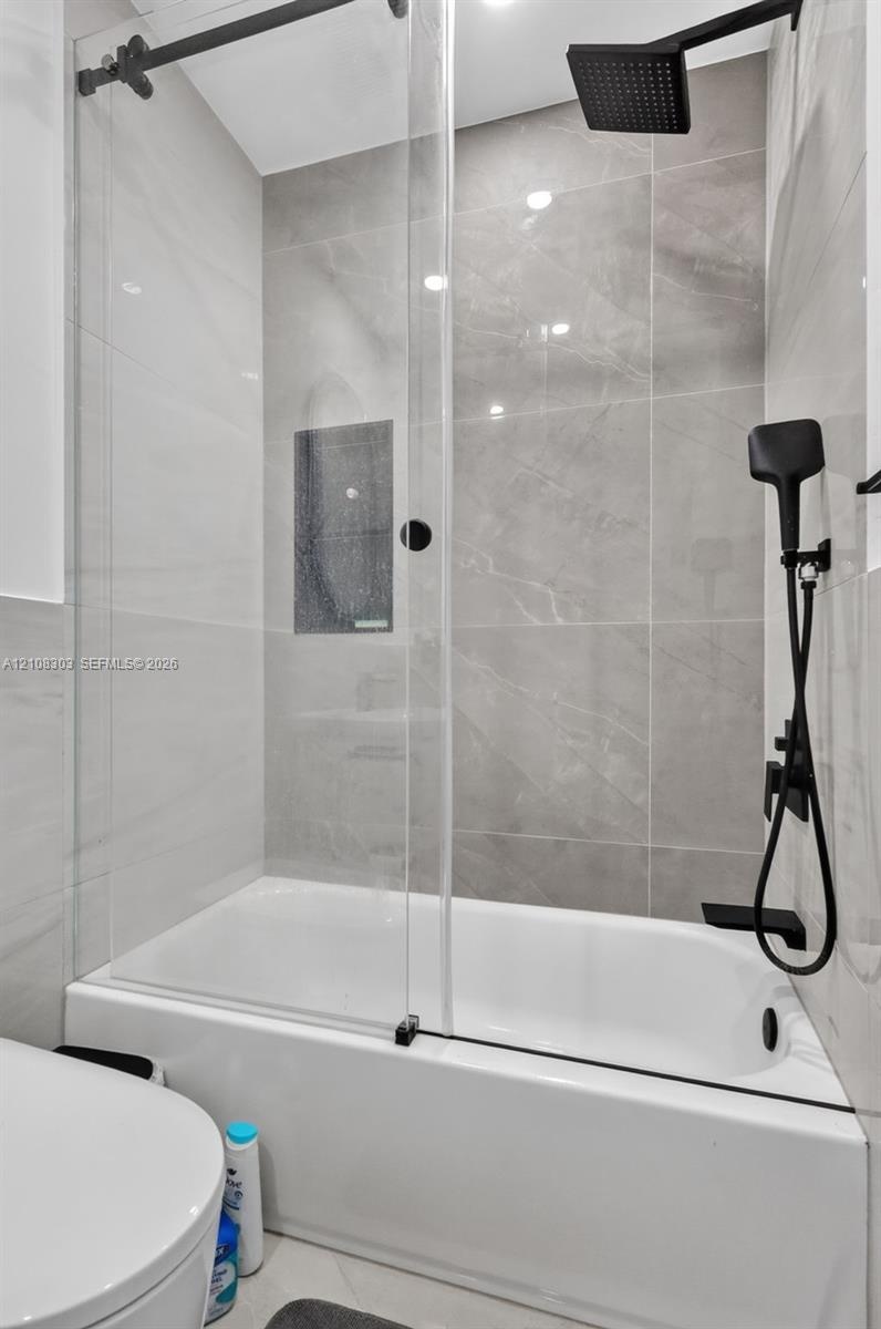 Shower and bathtub combination featuring large-format stone-finish tile surround, glass panel enclosure, matte black rainfall showerhead, and matching