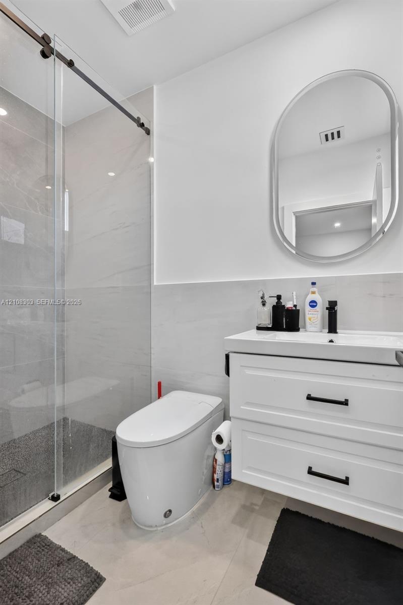 Walk-in shower with a sliding glass door, white wall-mounted vanity with two drawers, matte black hardware, oval-framed mirror, and large-format tile 