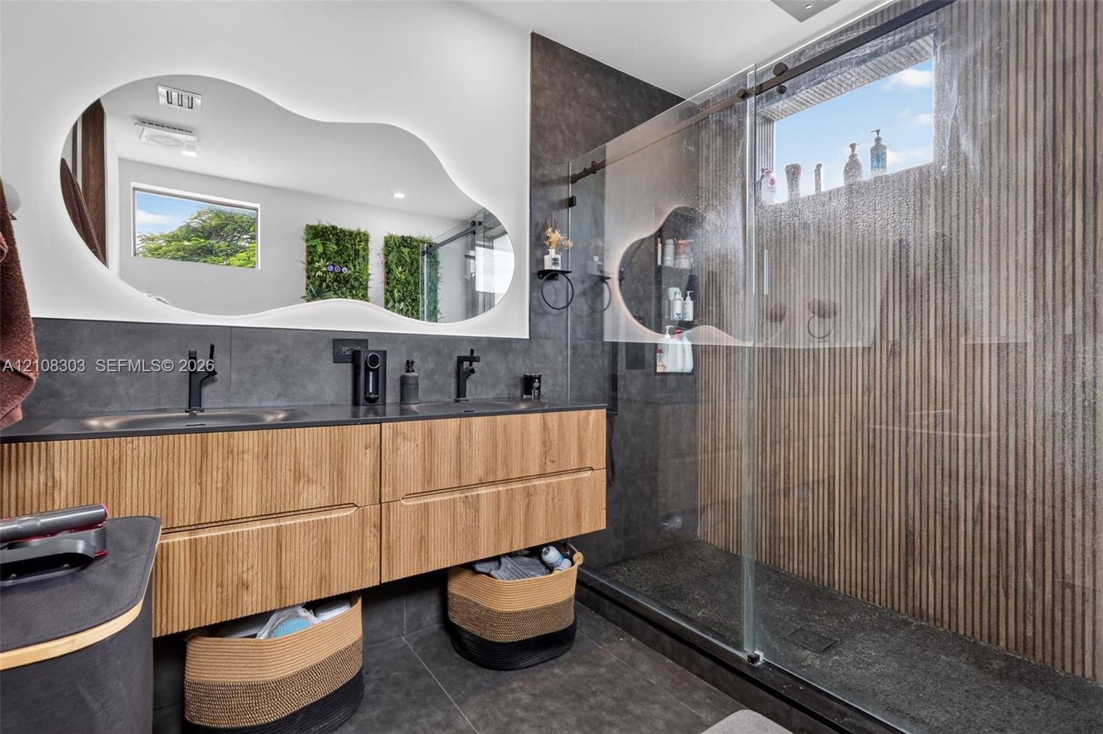 Floating wood-finish vanity with dual sinks, backlit organic-shaped mirror, matte black fixtures, walk-in shower with glass enclosure, and vertical-sl