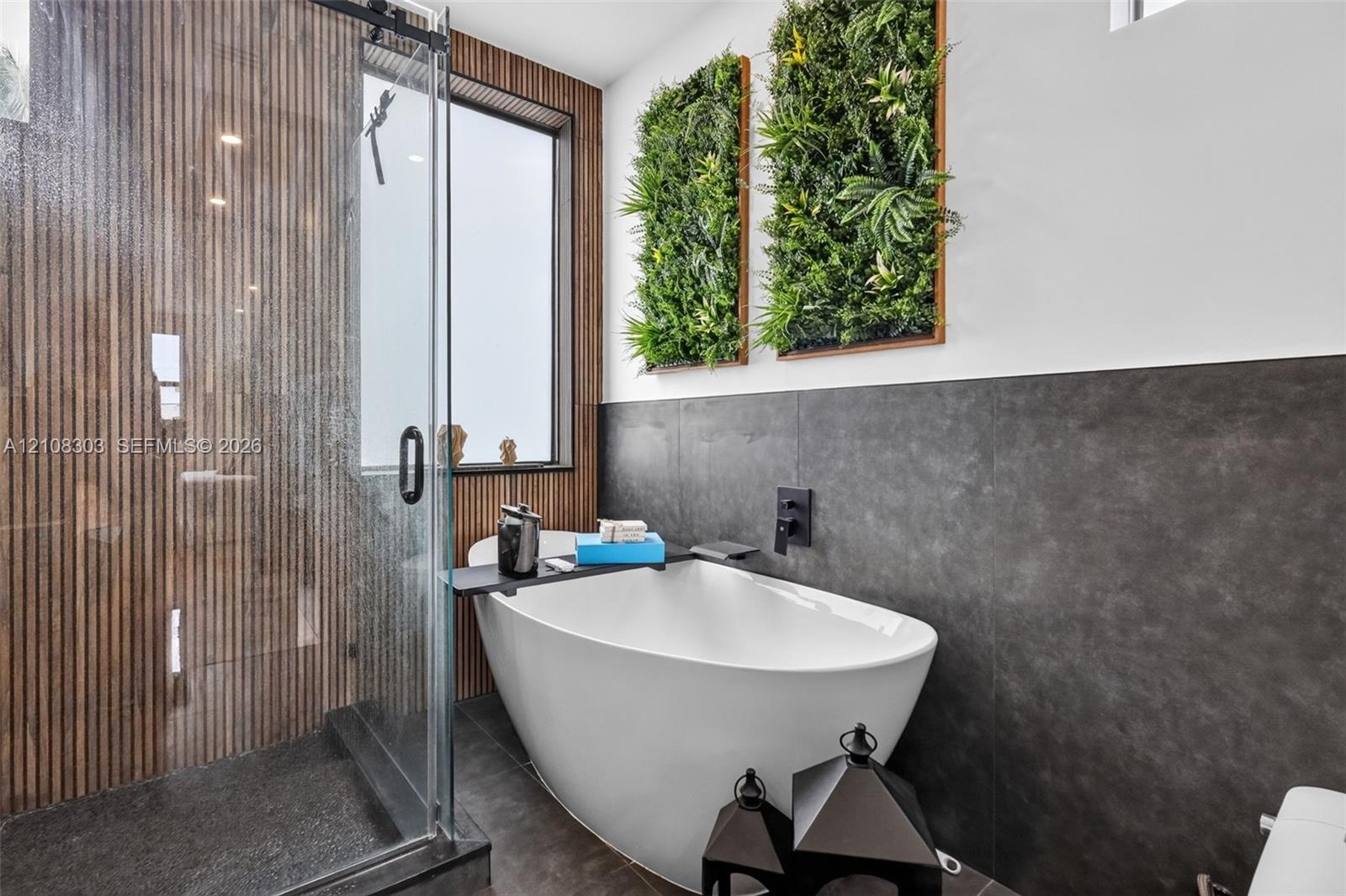 Freestanding soaking tub featuring matte black wall-mounted fixtures, vertical wood-slat accent wall, glass-enclosed shower, and large-format tile wal