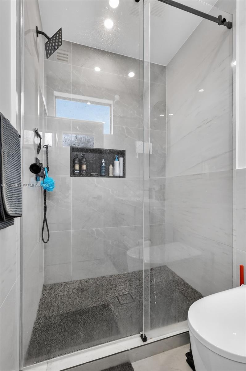 Walk-in shower featuring large-format light grey wall tile, a textured pebble-style floor, a built-in recessed niche, a frameless glass enclosure, and