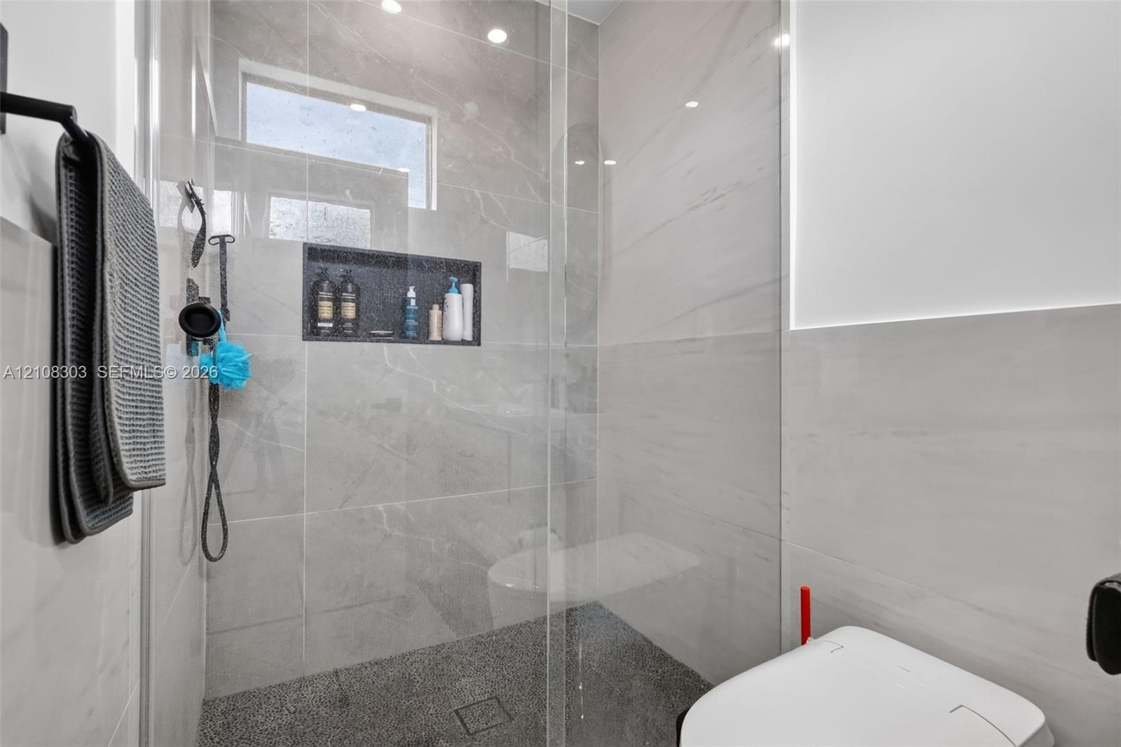 Walk-in shower featuring large-format stone-finish tile surround, integrated recessed storage niche, pebble-textured flooring, frameless glass enclosu
