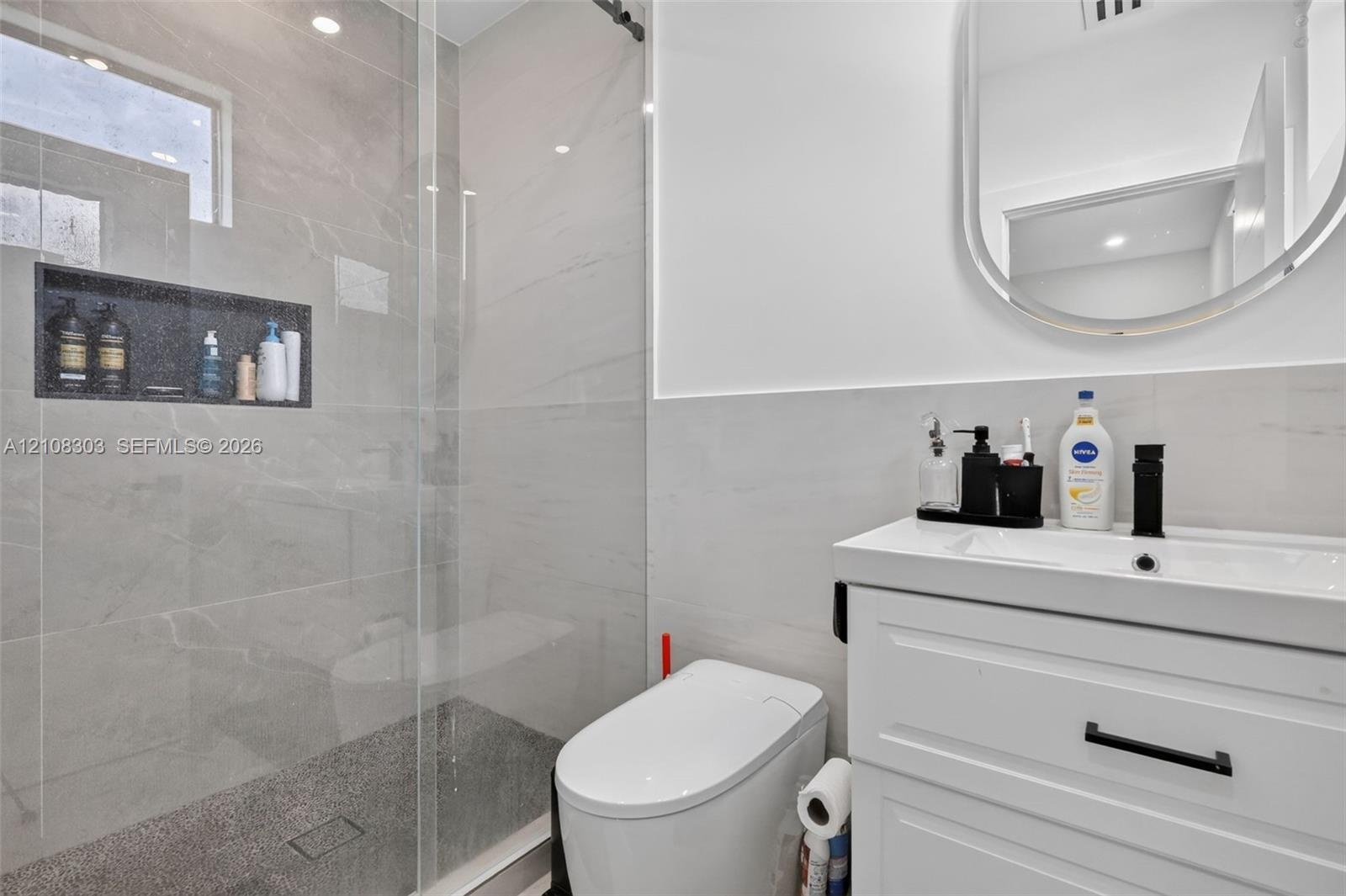 Walk-in shower with frameless glass enclosure, built-in tiled niche, white vanity with rectangular sink, matte black faucet, and oval wall-mounted mir