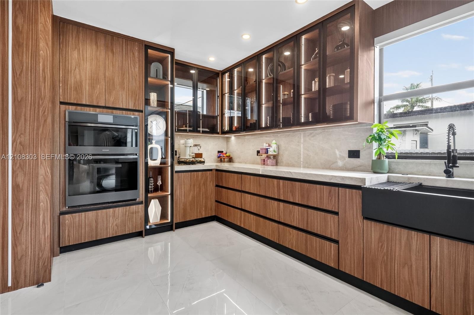 Wood-finish cabinetry with glass-front display panels, integrated double wall ovens, stone-finish countertops, matte black apron-front sink, and polis