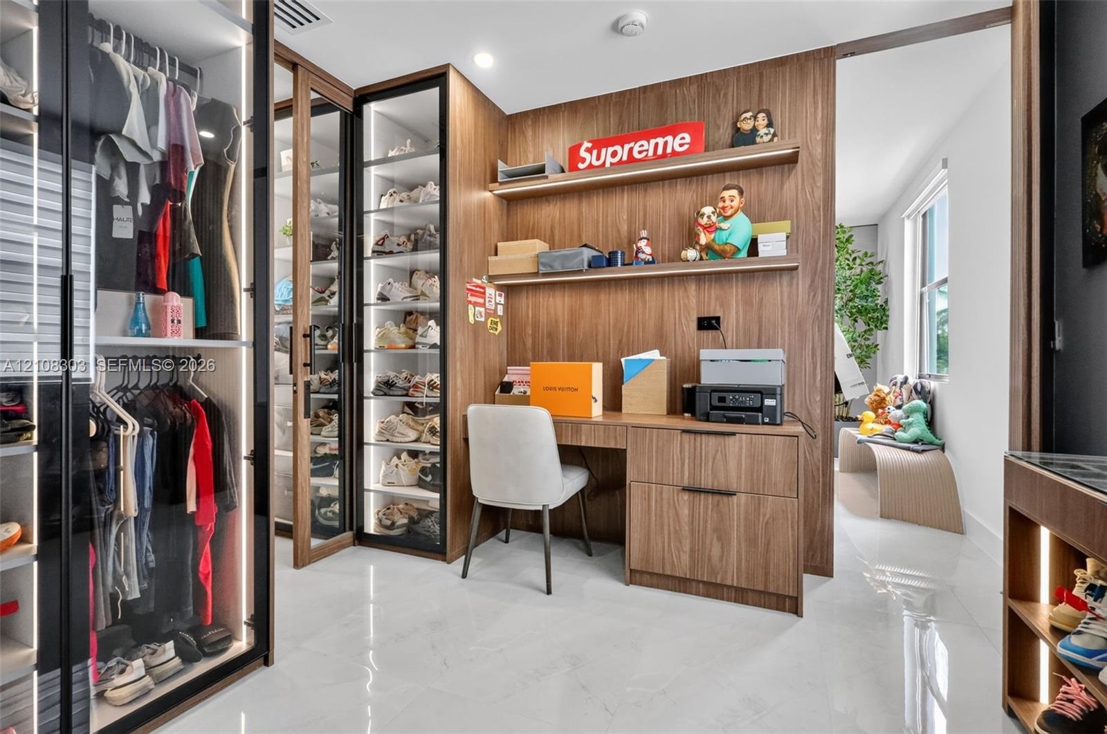 Custom dressing area featuring glass-front cabinetry with integrated lighting, wood-finish wall paneling, and a built-in workstation with floating she