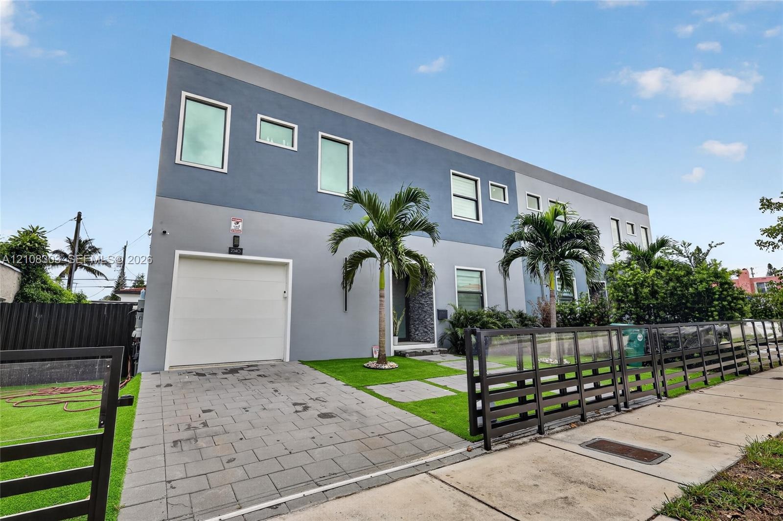 Contemporary two-story exterior featuring a smooth stucco finish, paver driveway, single-car garage, and integrated landscaping with mature palms