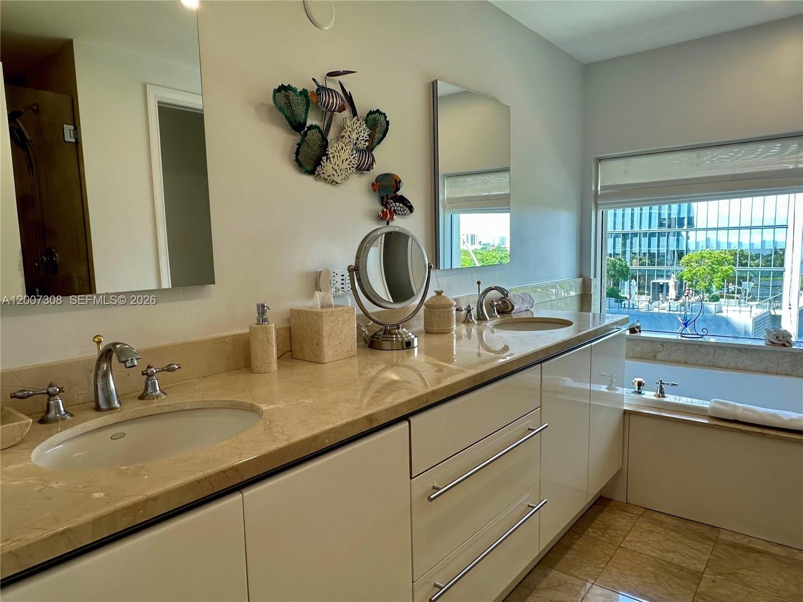 Dual-sink vanity with stone countertop, polished metal fixtures, flat-panel cabinetry, and inset soaking tub