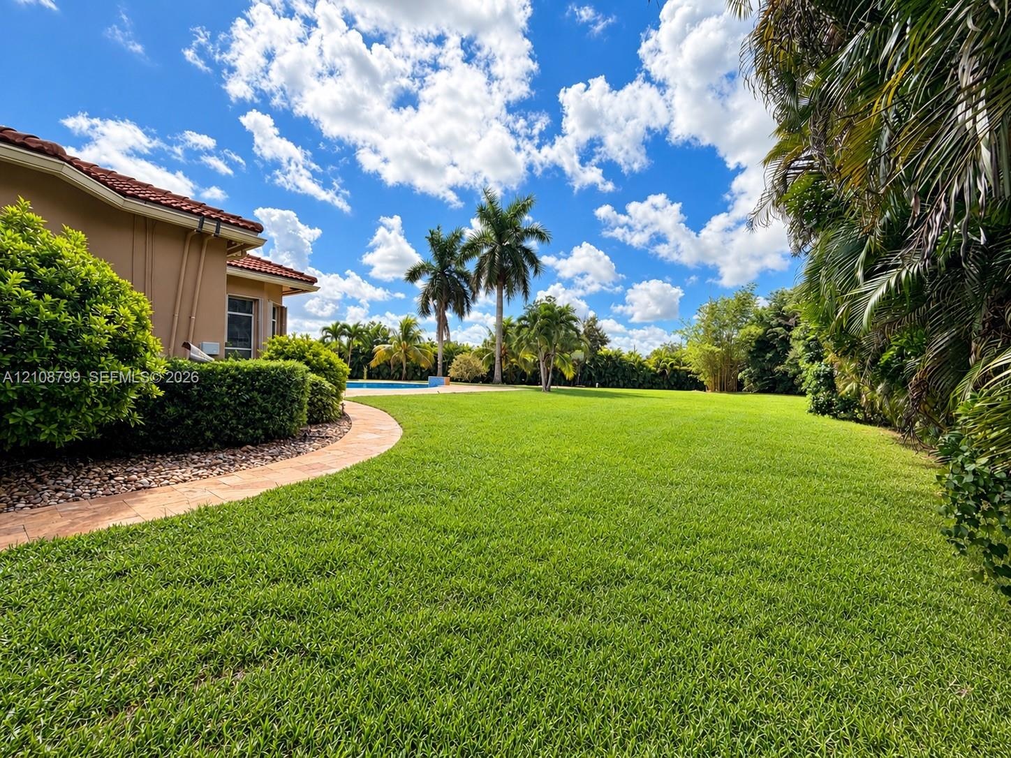 Expansive backyard featuring a manicured lawn, stone-paver walkway, swimming pool, and mature palm landscaping