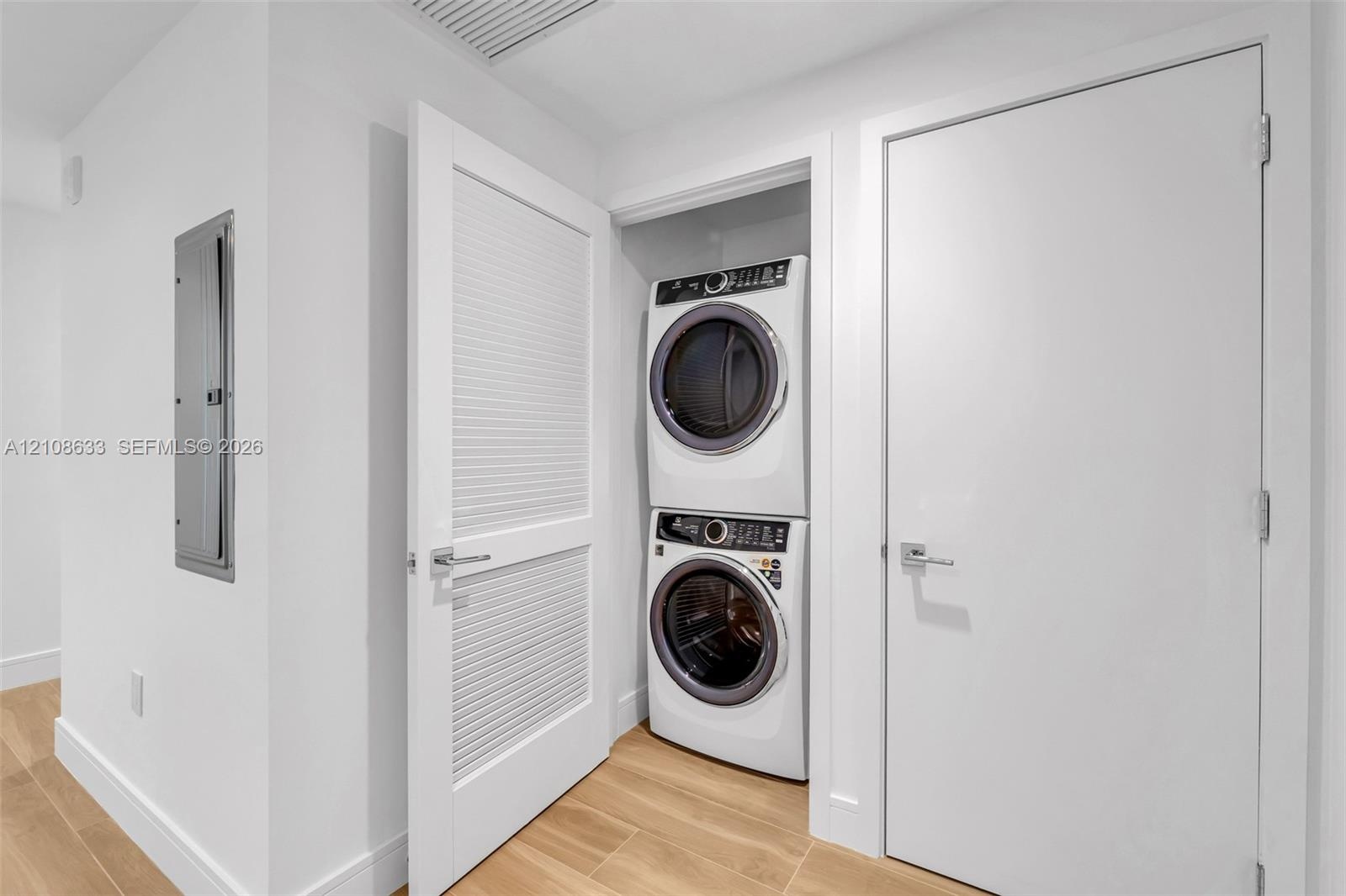 Laundry area featuring stacked front-load washer and dryer, wood-finish flooring, louvered closet door, and interior door with brushed nickel hardware