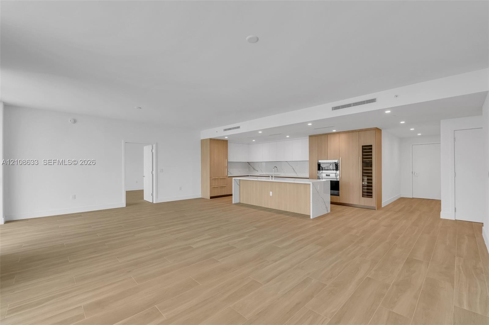 Kitchen featuring wood-finish cabinetry, an expansive waterfall-edge island, stone-surfaced backsplash, integrated wall oven, and wood-finish flooring