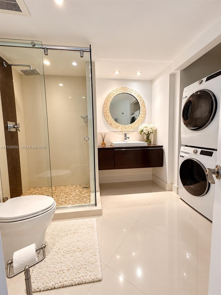 Glass-enclosed shower with a pebble-stone floor, floating dark-wood vanity, rectangular vessel sink, polished tile flooring, and integrated laundry ar