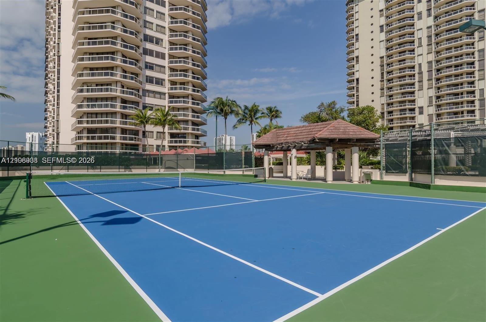 Tennis court featuring a blue surface with white boundary markings, perimeter fencing, and an open-air pavilion with a tiled roof