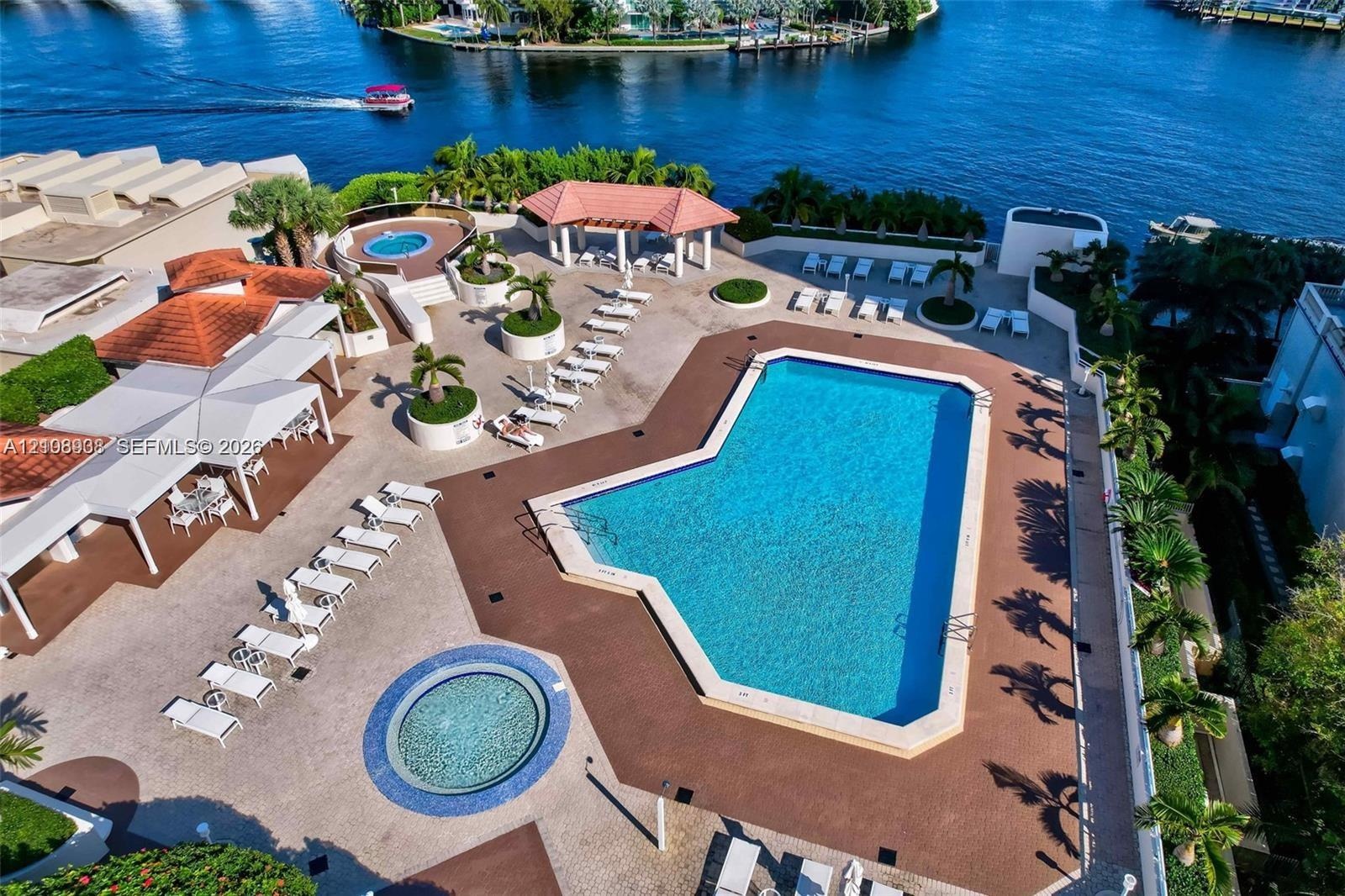Waterfront pool deck featuring an expansive pool, a circular spa, a covered pavilion with wood-beam construction, and extensive concrete hardscaping