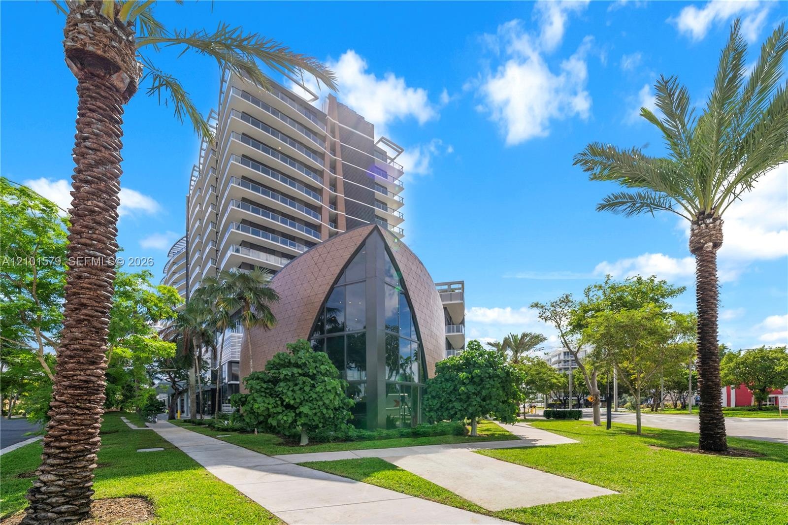 Multi-story architectural exterior featuring a curved metallic-toned facade, floor-to-ceiling glass paneling, tiered balconies, and manicured grounds 