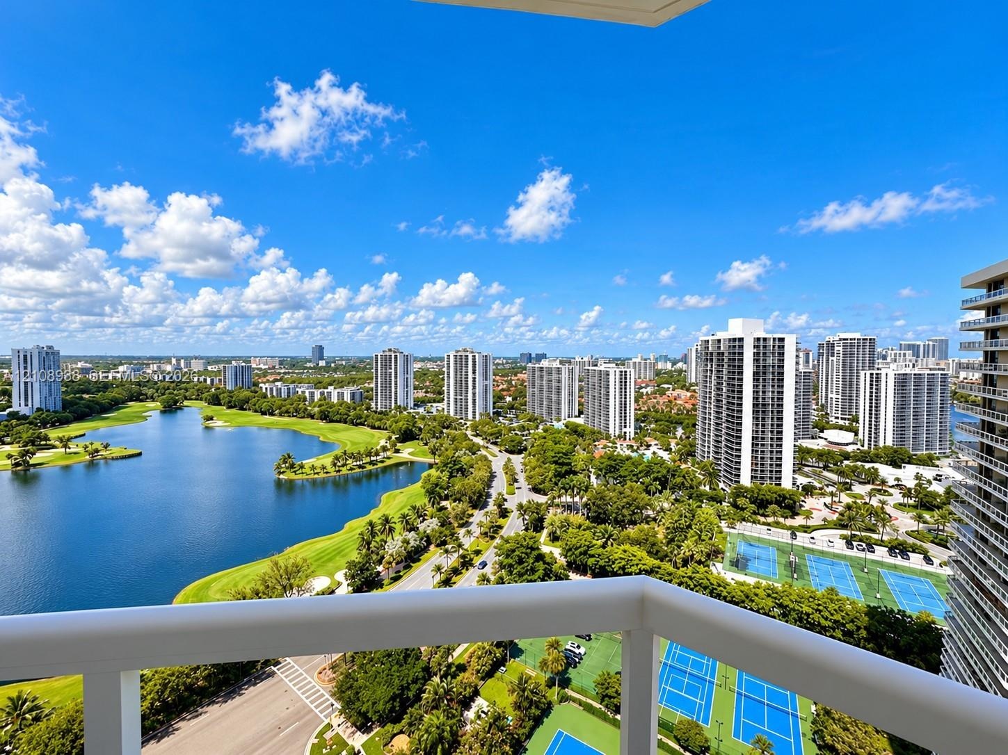 Elevated balcony featuring a white railing, overlooking an expansive lake, golf course fairways, and tennis courts