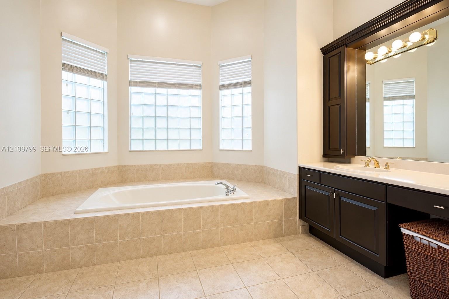 Soaking tub set in a tiled deck, bay window configuration with glass block accents, dark wood-finish cabinetry, countertop with integrated sink, and w