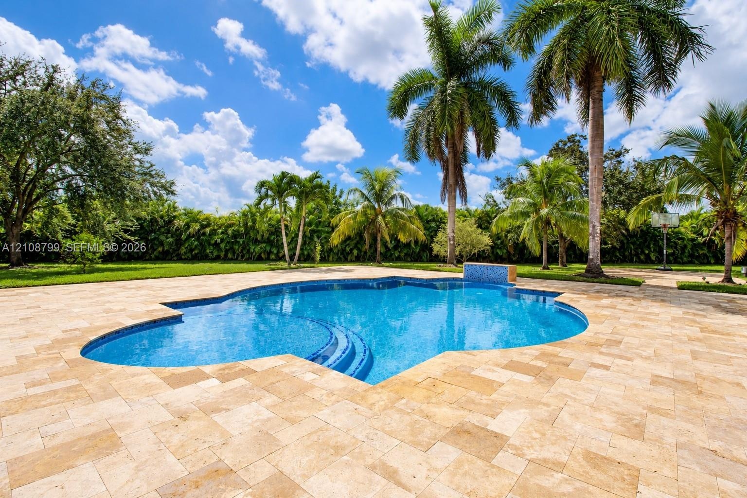 Free-form swimming pool featuring a tiled spillover water feature, stepped entry, and expansive stone-paver decking surrounded by established palm tre