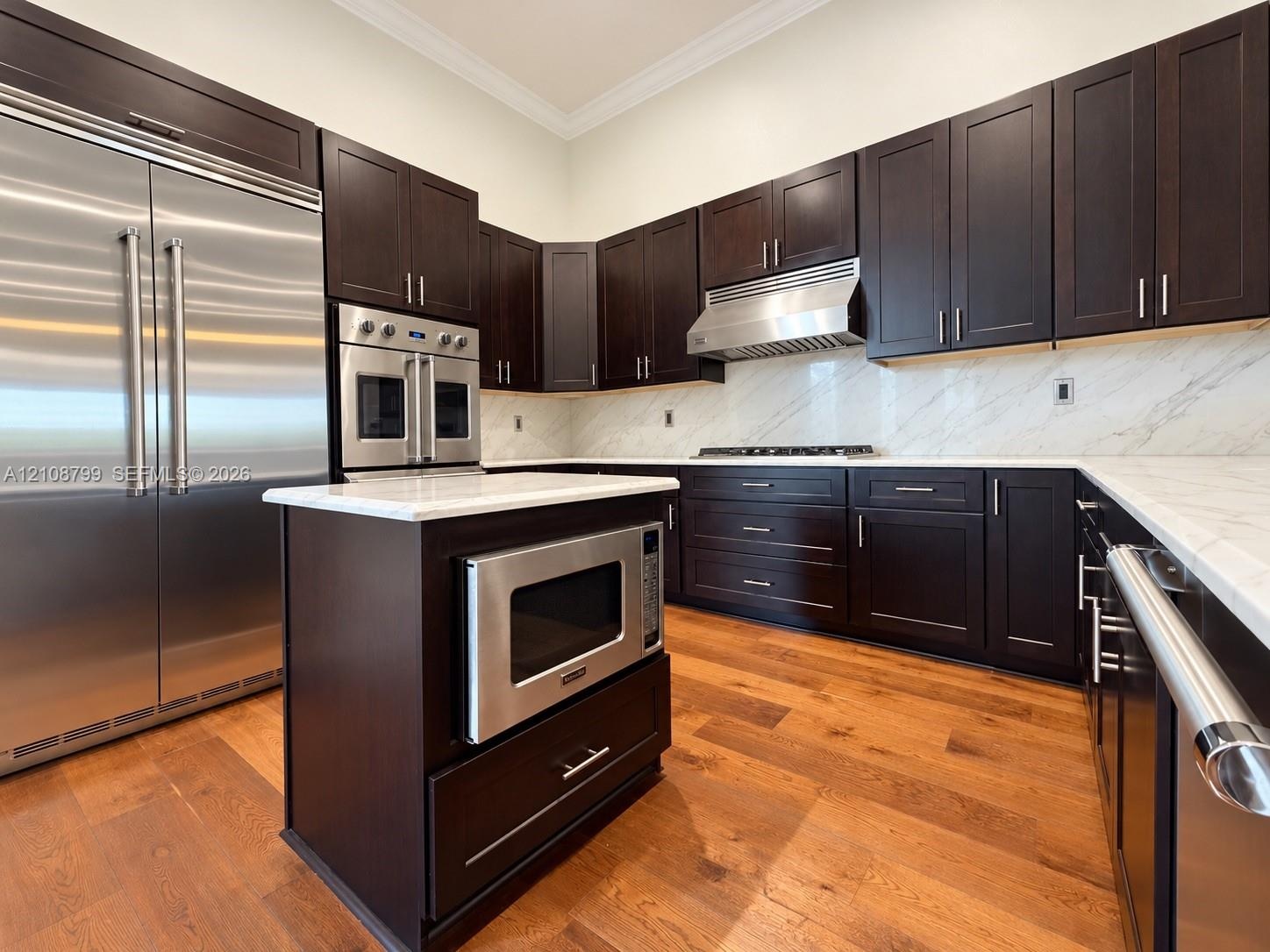 Kitchen featuring dark-finish cabinetry, stone-style countertops, stainless steel appliances, a center island with built-in microwave, and wood-finish