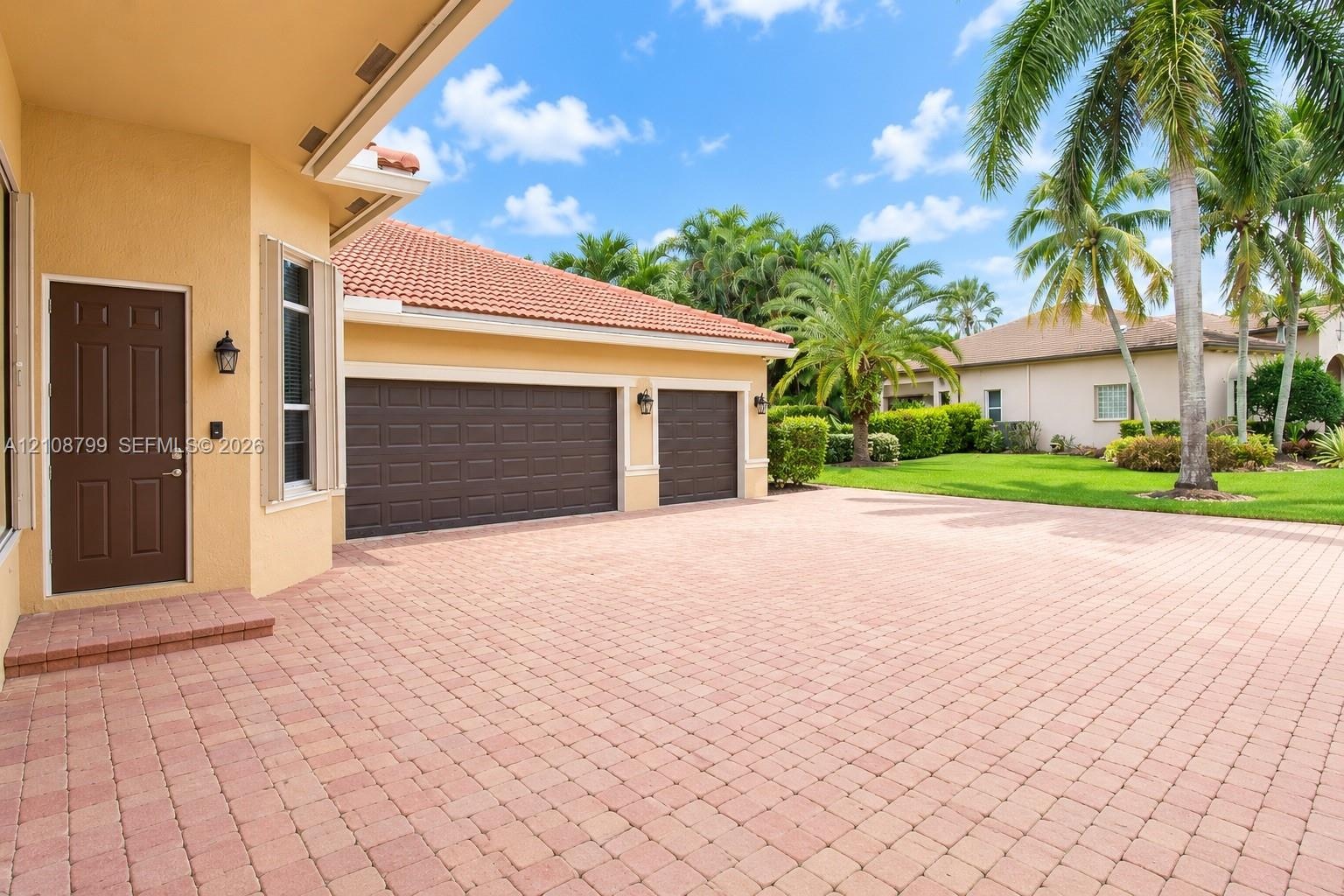 Paver driveway, three-car garage, stucco exterior, clay tile roof, and mature palm landscaping