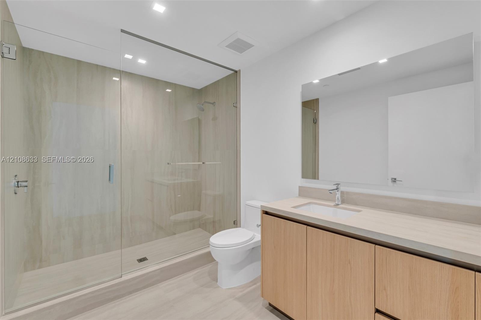 Walk-in shower with floor-to-ceiling neutral tile and frameless glass door, vanity featuring wood-finish cabinetry, solid-surface countertop with inte