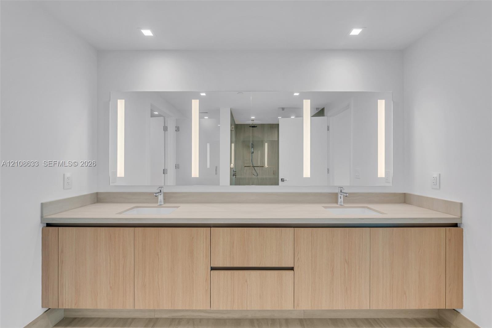 Floating wood-finish vanity with dual integrated sinks, light-toned stone countertop, expansive wall-to-wall mirror with vertical integrated lighting,
