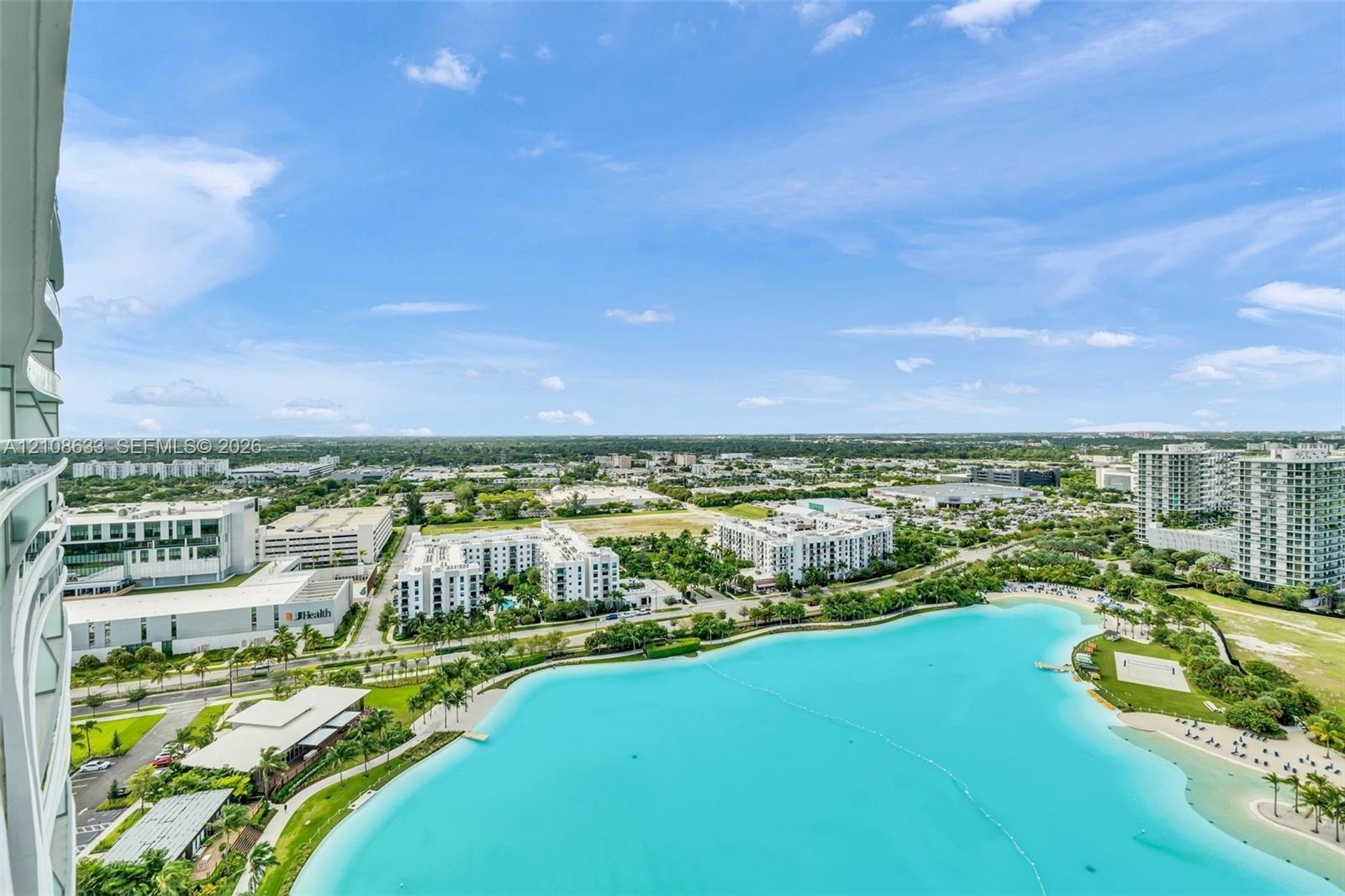 Elevated balcony featuring a glass panel railing, showcasing a panoramic perspective of a large turquoise lagoon, sandy beach area, and surrounding la