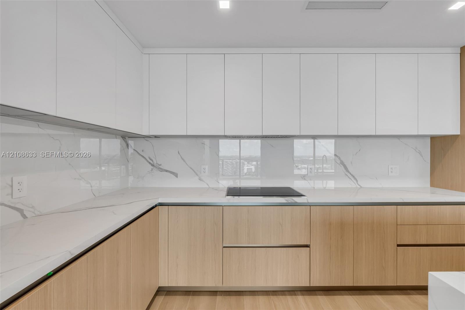 Contemporary kitchen featuring flat-panel upper cabinetry, light-toned wood base cabinetry, stone-surface countertops, full-height stone-surface backs