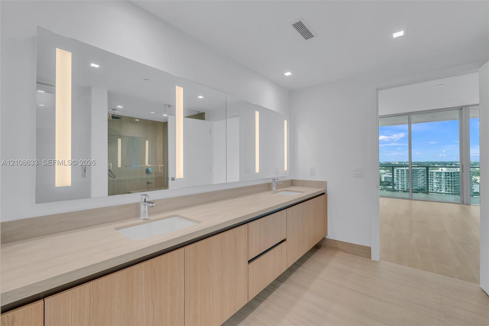 Floating vanity with integrated dual rectangular sinks, light wood-finish cabinetry, full-length illuminated wall mirror, and neutral-tone flooring