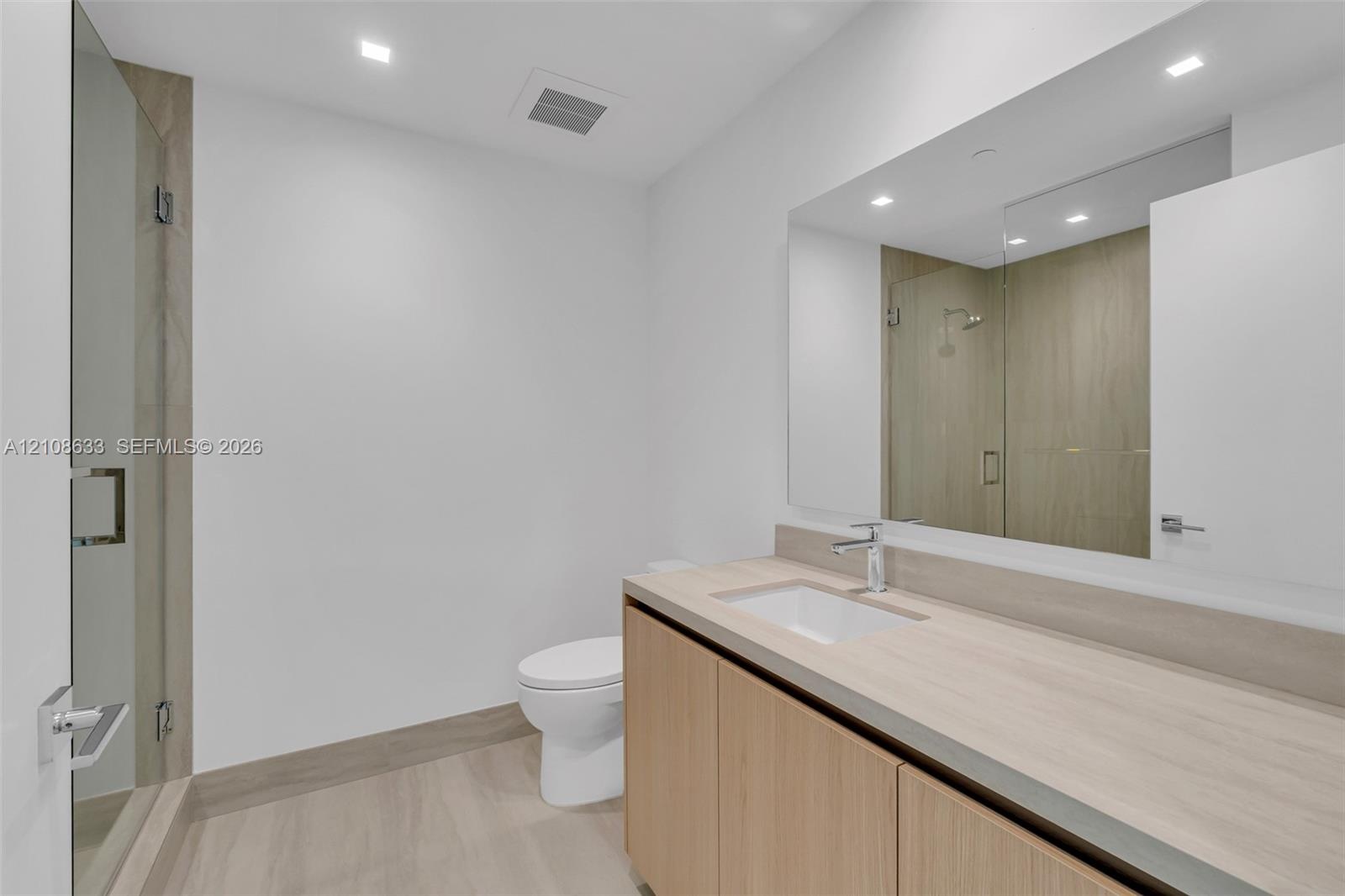 Floating vanity with light wood-finish cabinetry, integrated rectangular sink, large wall-spanning mirror, and glass-enclosed shower with neutral-tone