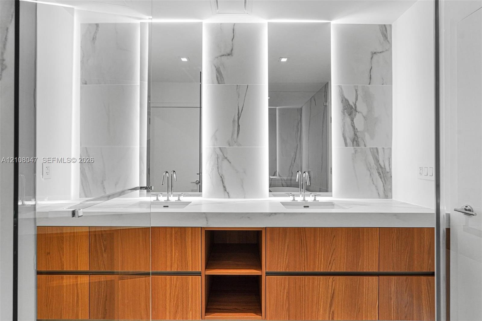 Dual vanity featuring wood-finish cabinetry, integrated sinks, a solid surface countertop, and backlit mirrors with wall-mounted stone-look tiles