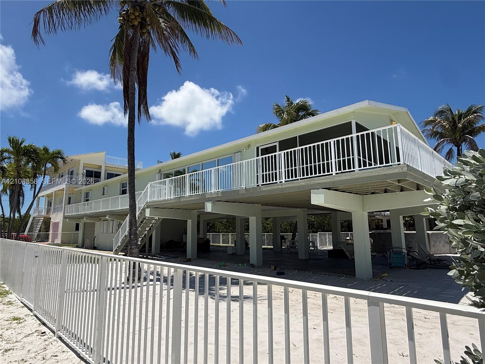 Elevated exterior featuring white metal railings, a screened balcony, concrete pilings, and a sandy ground-level area