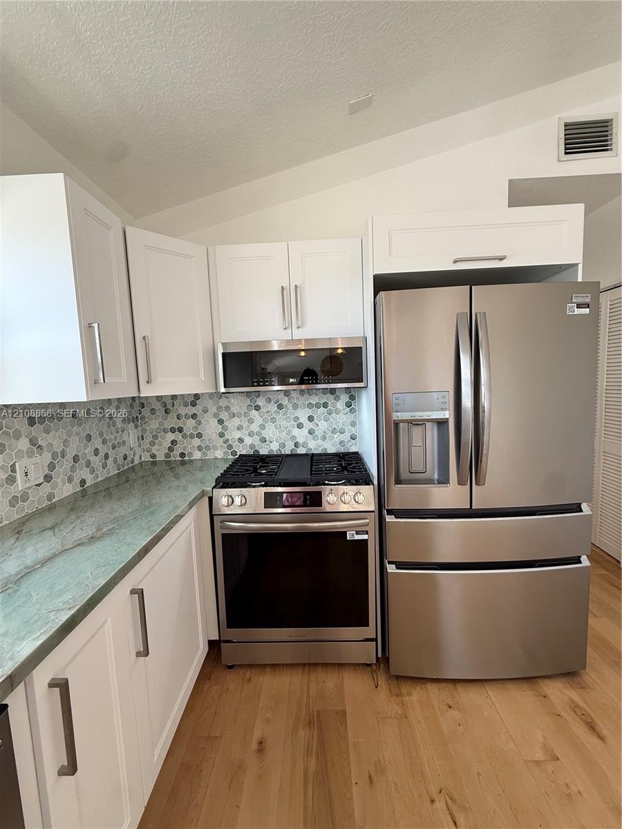 Kitchen featuring white shaker-style cabinetry, natural stone countertops, hexagonal tile backsplash, stainless steel appliances, and wood-finish floo