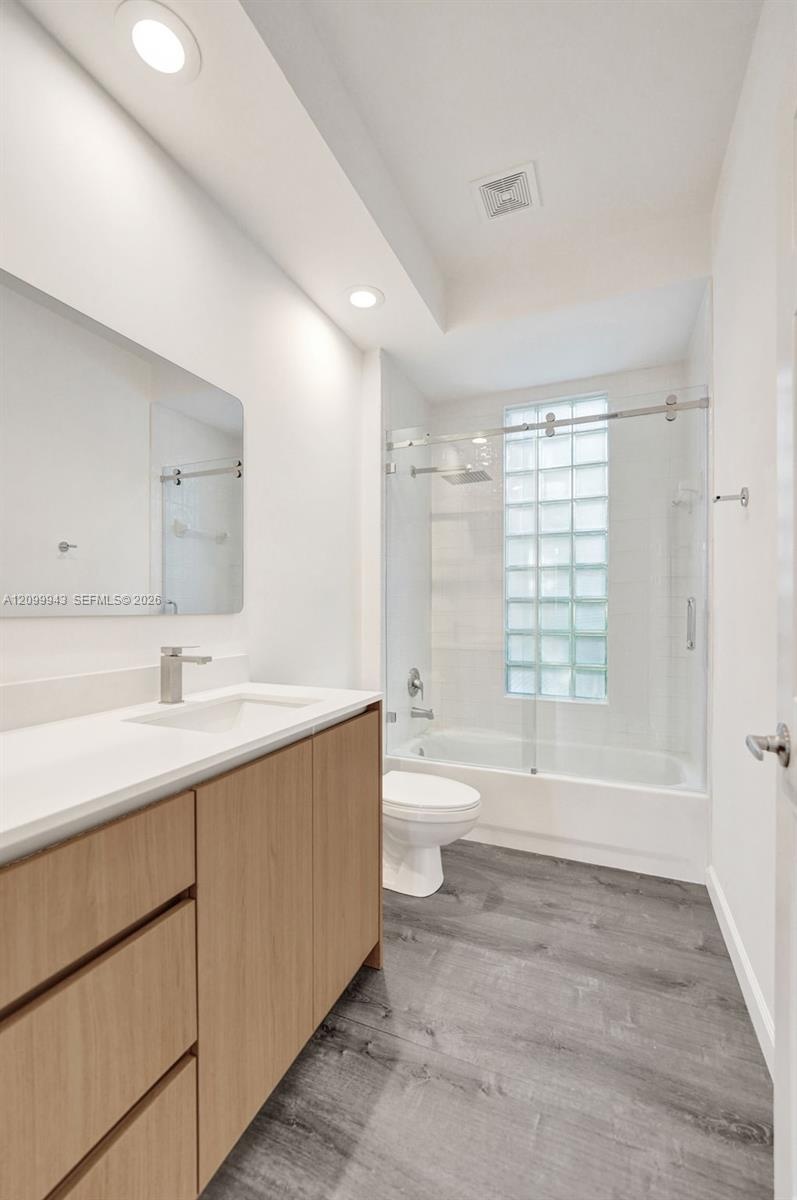 Wood-finish cabinetry with a solid surface countertop, recessed lighting, glass block window, sliding glass shower door, and gray wood-finish flooring