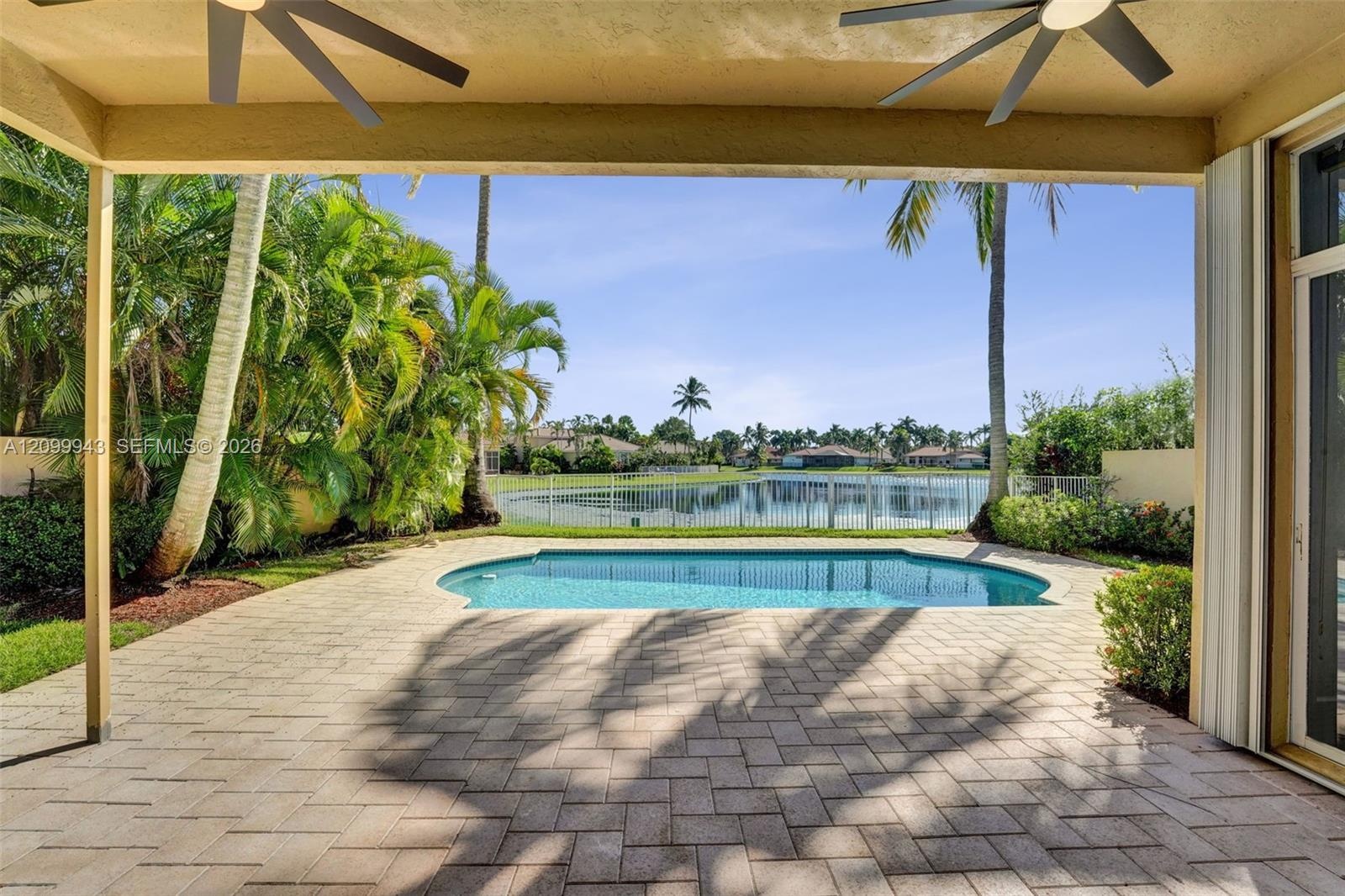 Covered patio featuring paver flooring and ceiling fans, opening to a pool with a metal perimeter fence and mature palm trees