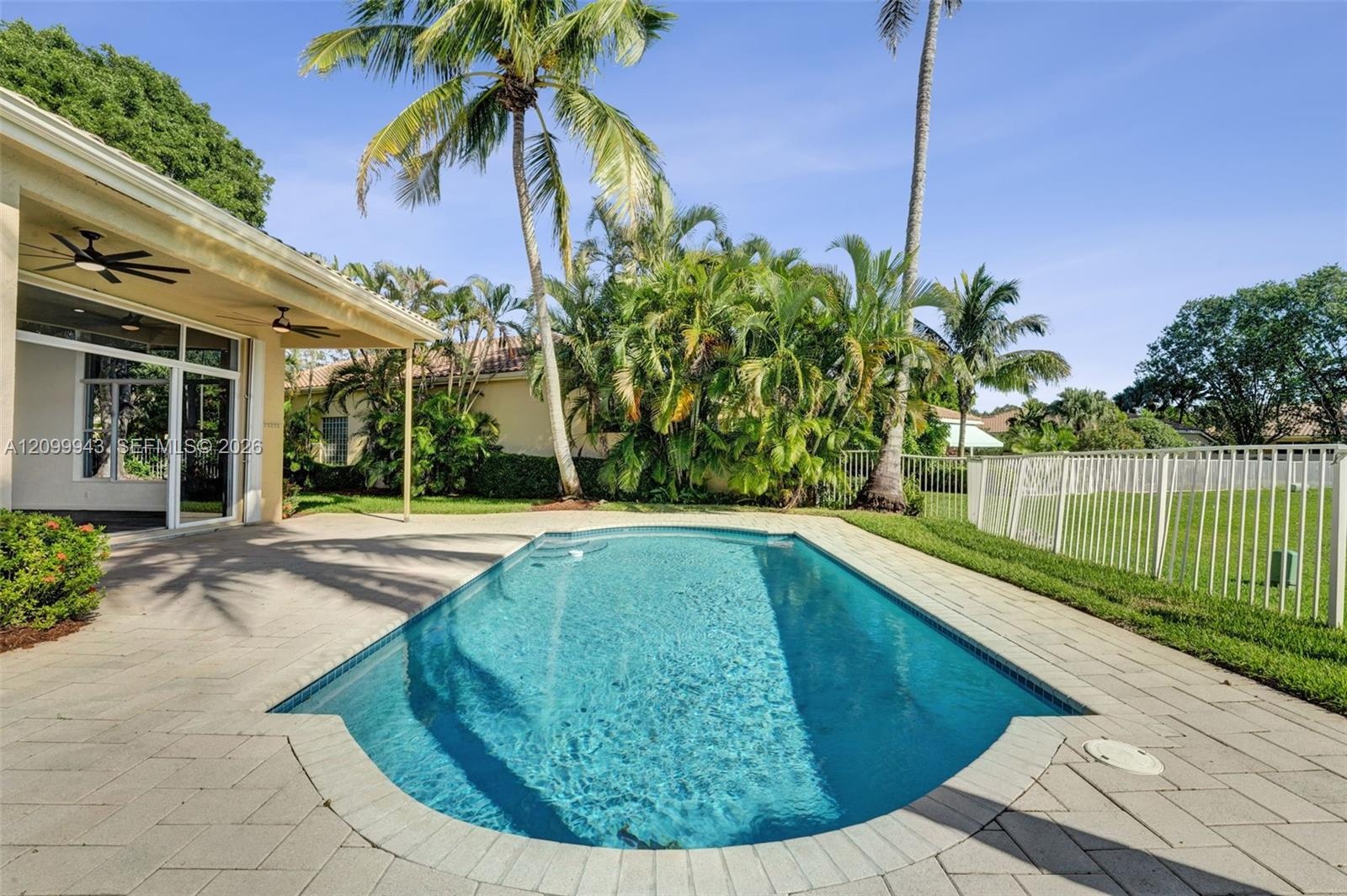 In-ground swimming pool with a stone paver deck, covered patio, white metal fencing, and mature palm trees
