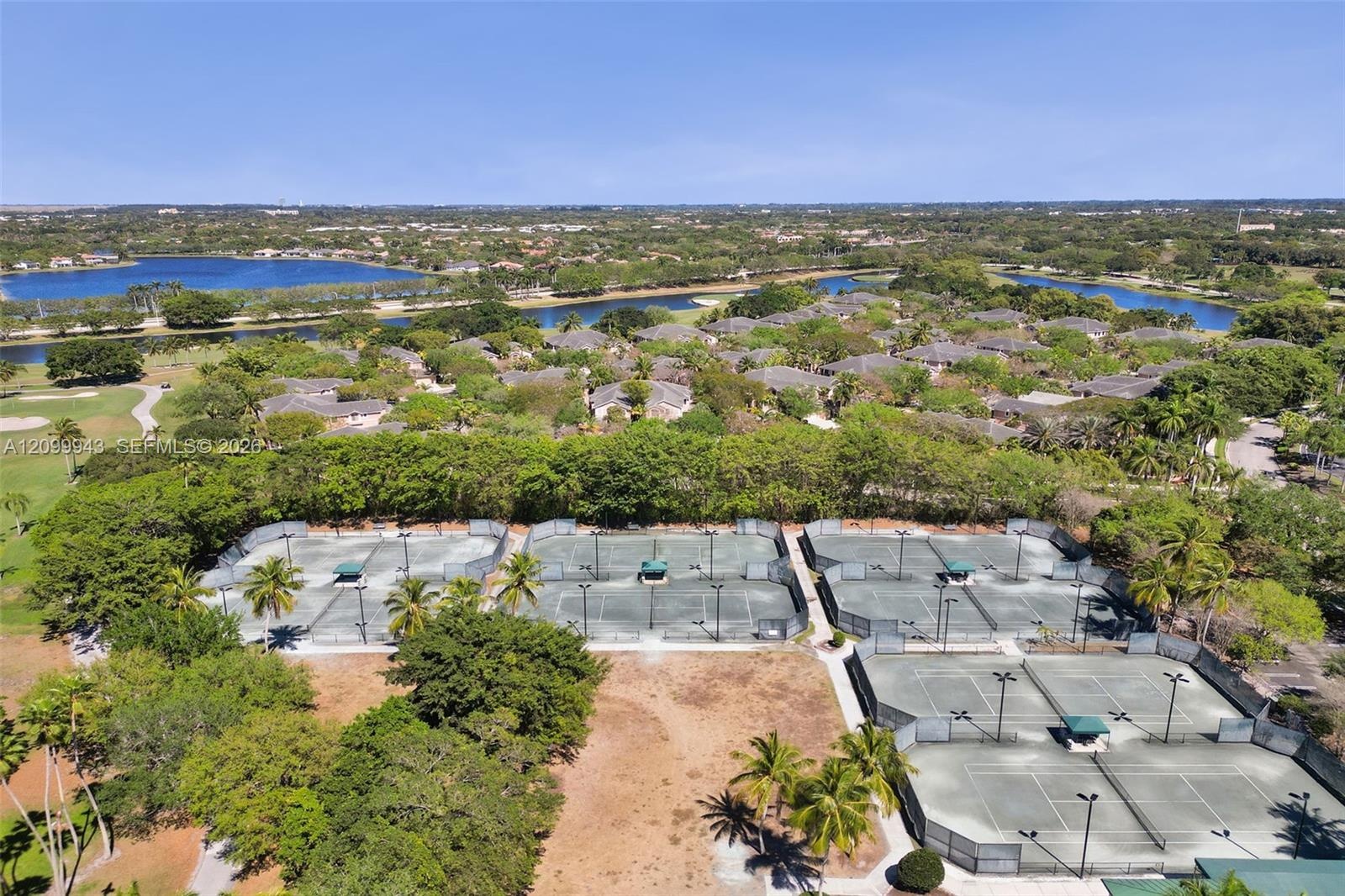 Community tennis complex featuring multiple hard-surface courts, perimeter fencing, overhead lighting, and shaded seating areas