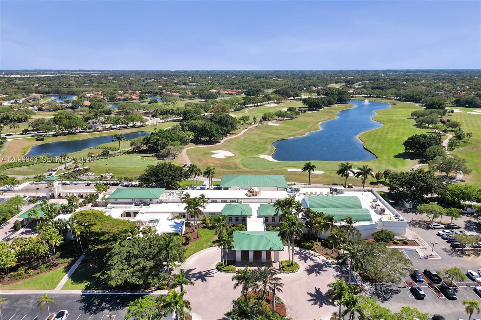 Expansive clubhouse facility featuring a multi-gabled green metal roof, a circular porte-cochère entry, mature palm tree landscaping, and paved parkin