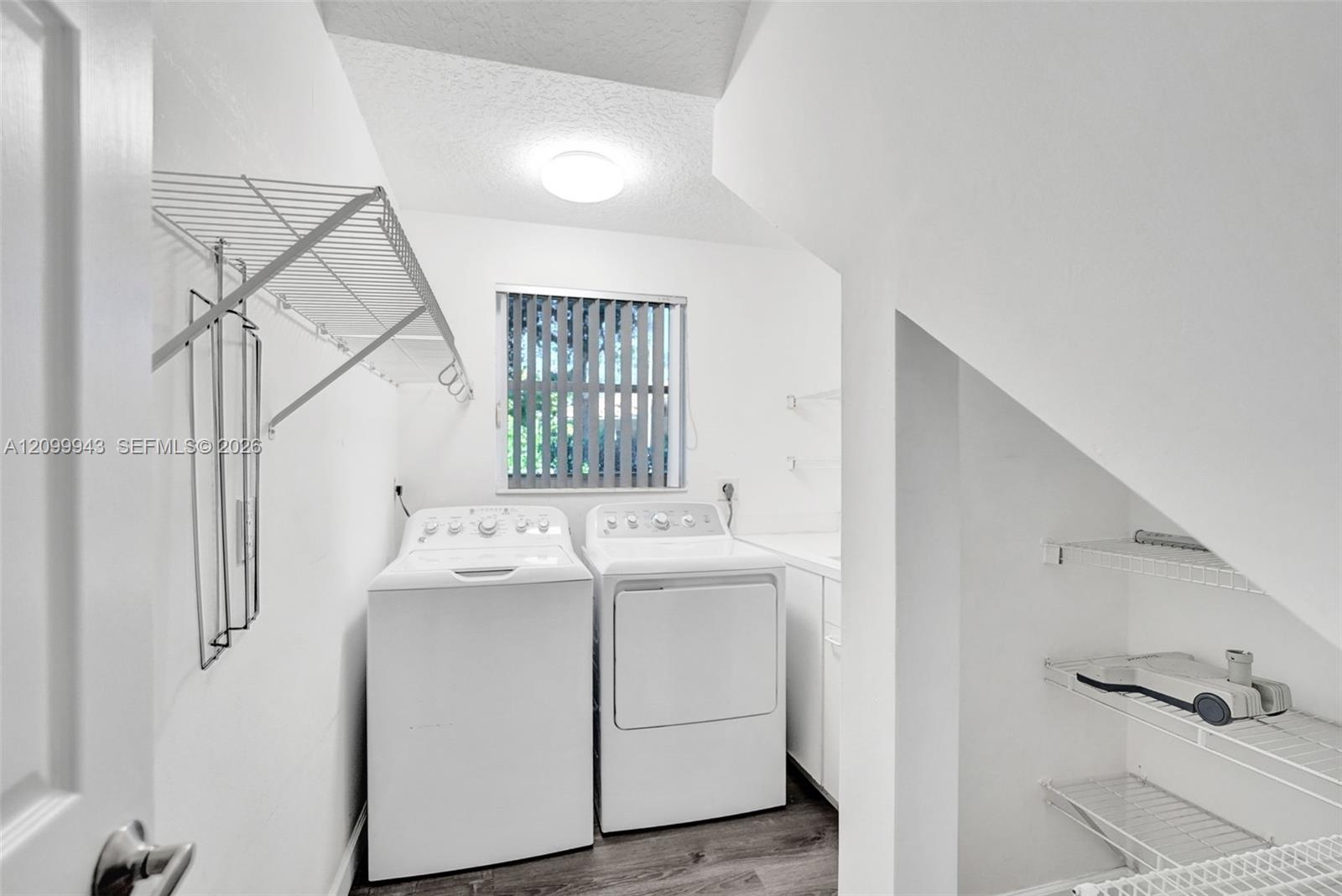 Laundry area featuring wood-finish flooring, white cabinetry, wire shelving, and vertical window blinds