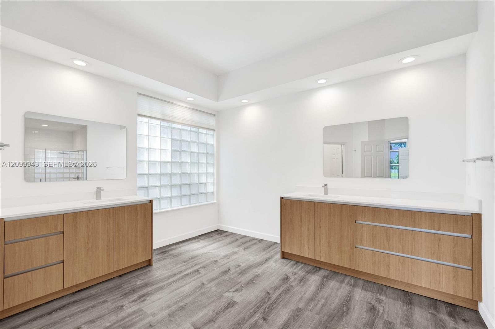 Dual wood-finish vanities with solid-surface countertops, integrated sinks, glass block window, and recessed ceiling lighting